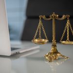 Balança dourada da justiça sobre uma mesa, ao lado de um computador portátil.