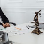 Mesa de trabalho jurídico com documentos, notebook e uma estátua da Justiça.